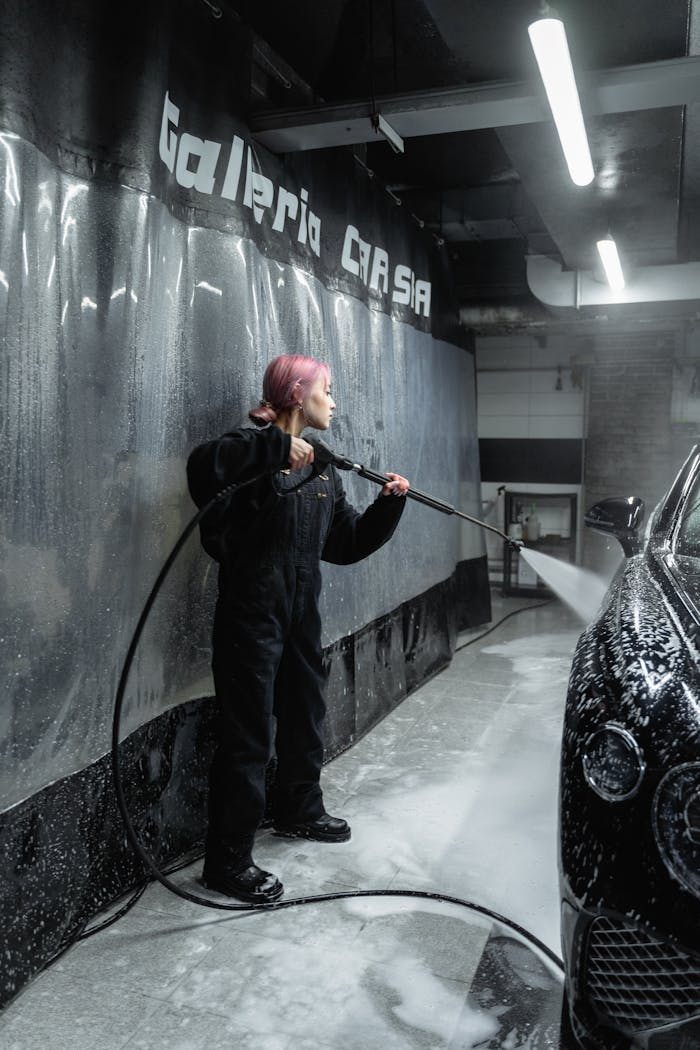 A young woman in uniform cleans a car with a power spray in an indoor garage setting.